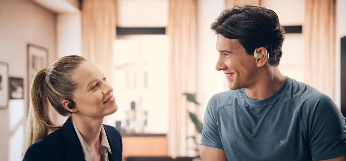 Kobieta i mężczyzna w słuchawkach uśmiechają się do siebie. Są w pokoju z naturalnym światłem. Tło ma zasłony.