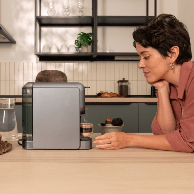 Kobieta w kuchni obserwuje parzenie kawy z srebrnej maszyny. Szklanka kawy stoi na blacie.