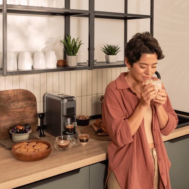 Kobieta w różowej koszuli pije kawę w kuchni. Ekspres do kawy, filiżanki i przekąski na blacie. Rośliny na półce.