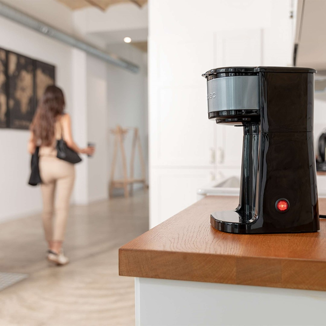 Czarny ekspres do kawy na drewnianym blacie. W tle odchodzi kobieta.