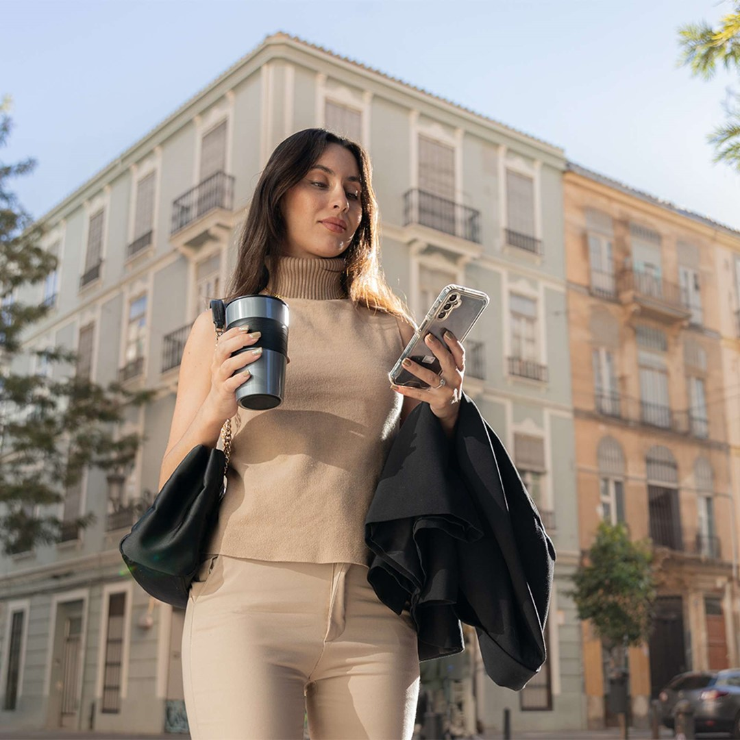 Kobieta trzymająca kubek z kawą i telefon, patrząca na telefon, stojąca przed budynkiem. Ma na sobie beżowe ubrania.
