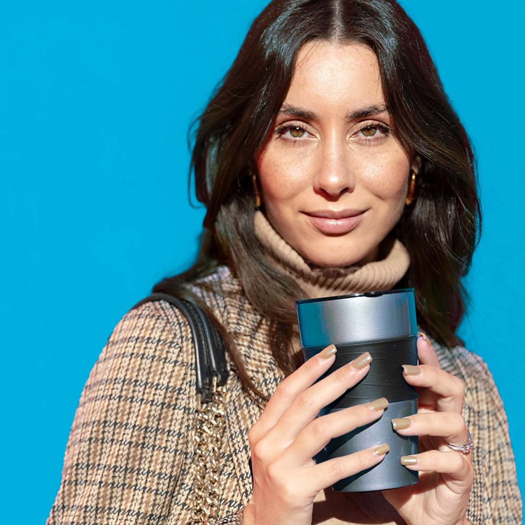 Kobieta z długimi brązowymi włosami trzyma szaro-czarny kubek. Ma na sobie beżowy golf i płaszcz w kratę na niebieskim tle.