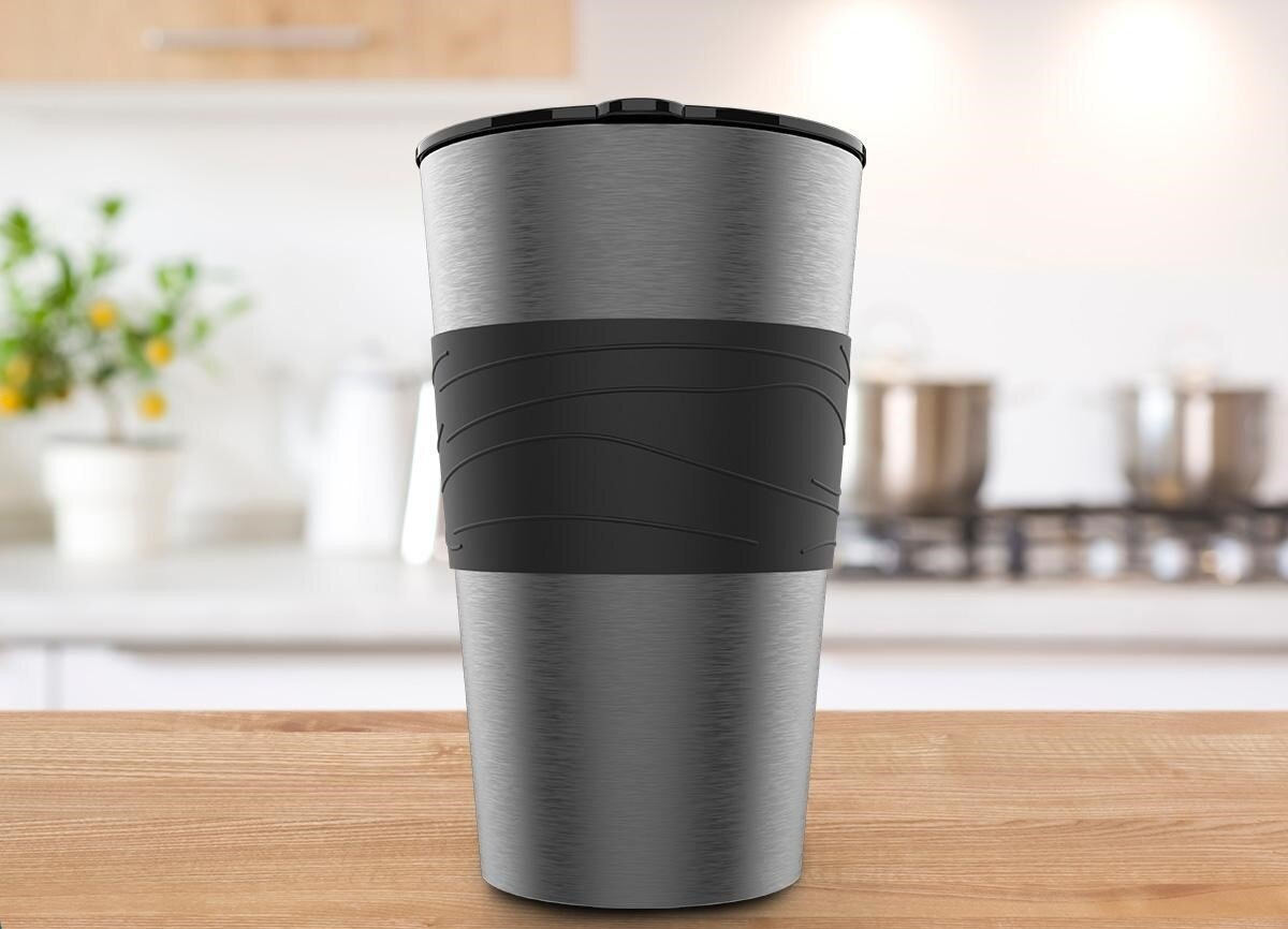 Srebrny kubek termiczny z czarną pokrywką i uchwytem. Stoi na drewnianym stole, z rozmytym tłem kuchni.