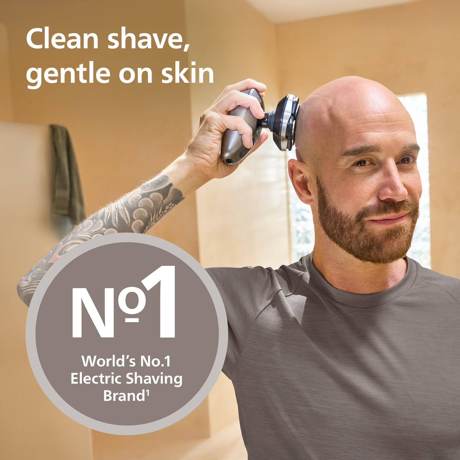 A bald man shaves his head with an electric shaver. He has a beard and a tattoo on his arm. The image promotes a shaving brand.