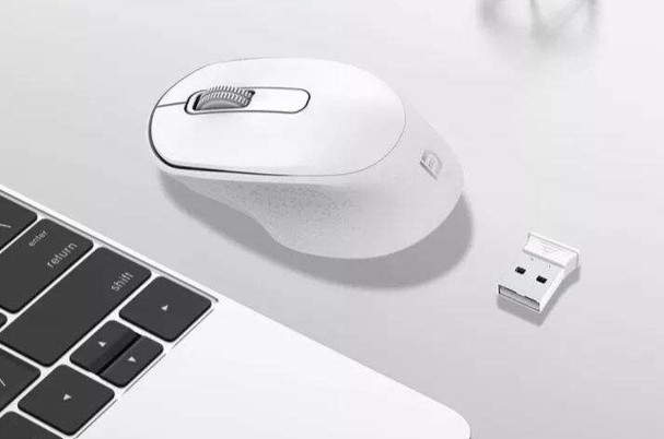 Beyaz bir kablosuz fare ve USB alıcısı, bir dizüstü bilgisayar klavyesinin yanında beyaz bir yüzeyde.