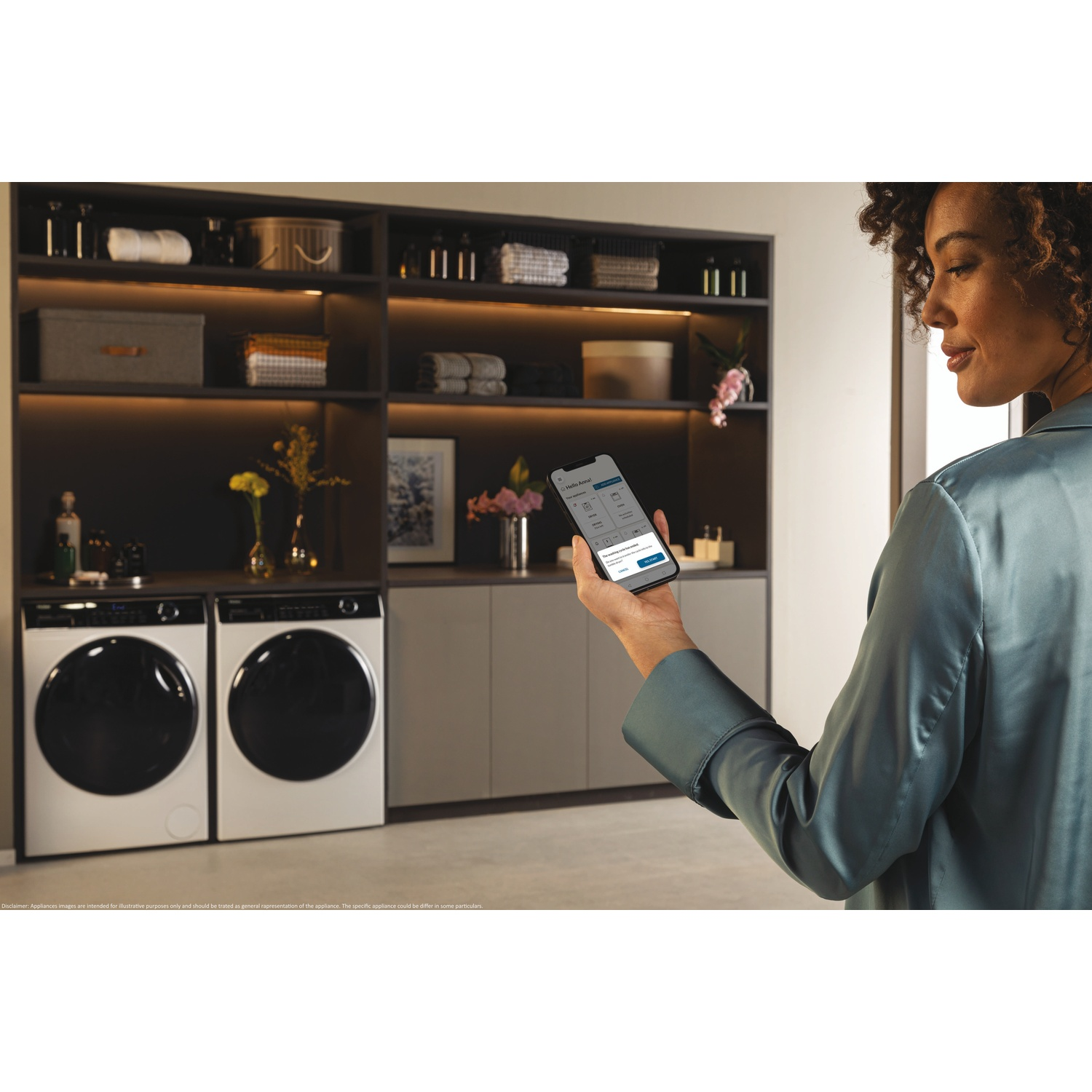 Donna che usa uno smartphone per controllare una lavatrice. Due lavatrici bianche in primo piano, sfondo è un mobile contenitore.