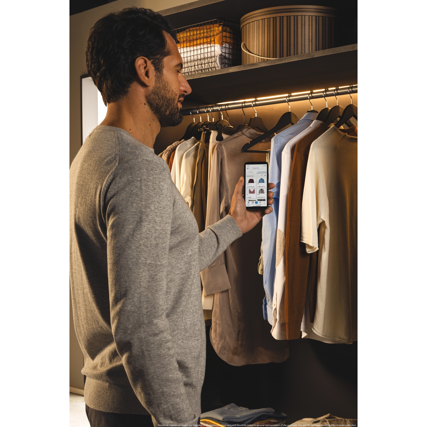 Un uomo in maglione grigio guarda il suo telefono che mostra vestiti. È davanti a un armadio con camicie appese e un cesto.