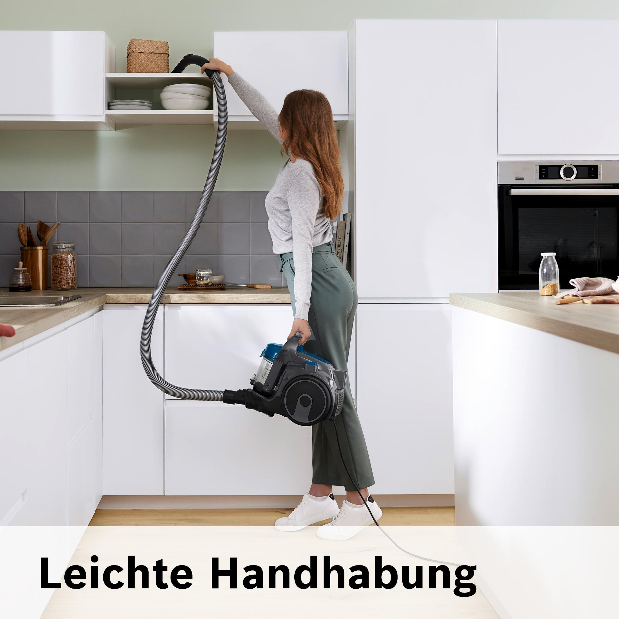 Eine Frau saugt eine Küche. Sie hält einen Staubsauger, der Schlauch reicht bis zu einem Schrank. Die Küche hat weiße Schränke.