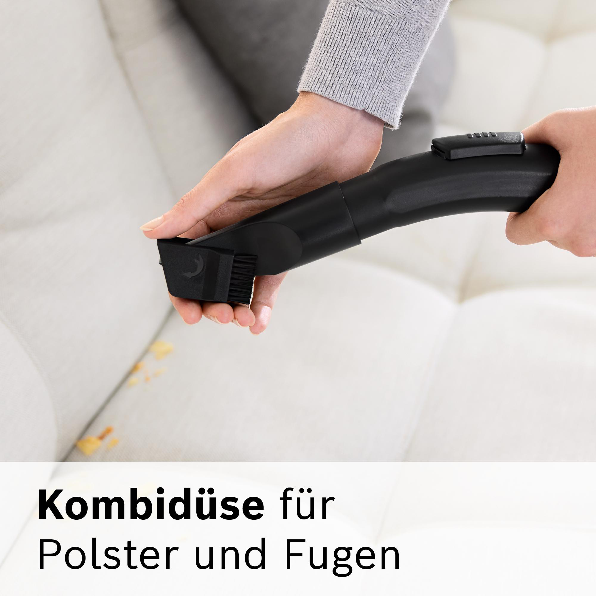 Eine Person benutzt einen schwarzen Staubsauger mit Polsterdüse auf einem weißen Sofa. Das Sofa hat gelbe Flecken.