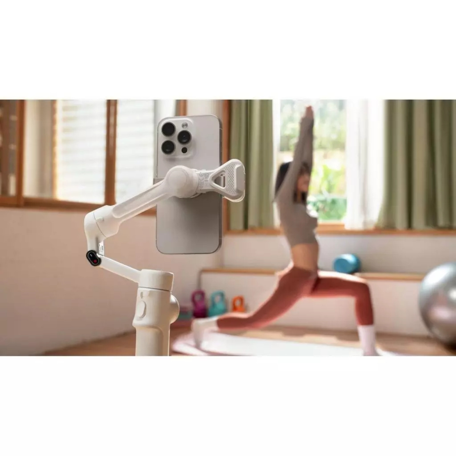 A woman in yoga pose, a phone on a stand is in front of her. The room has a window and yoga equipment.