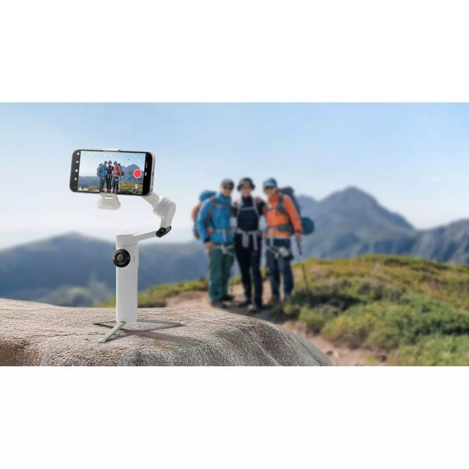 A phone records hikers with a gimbal. The phone shows other hikers, with mountains in the background. Gimbal is on a stone.