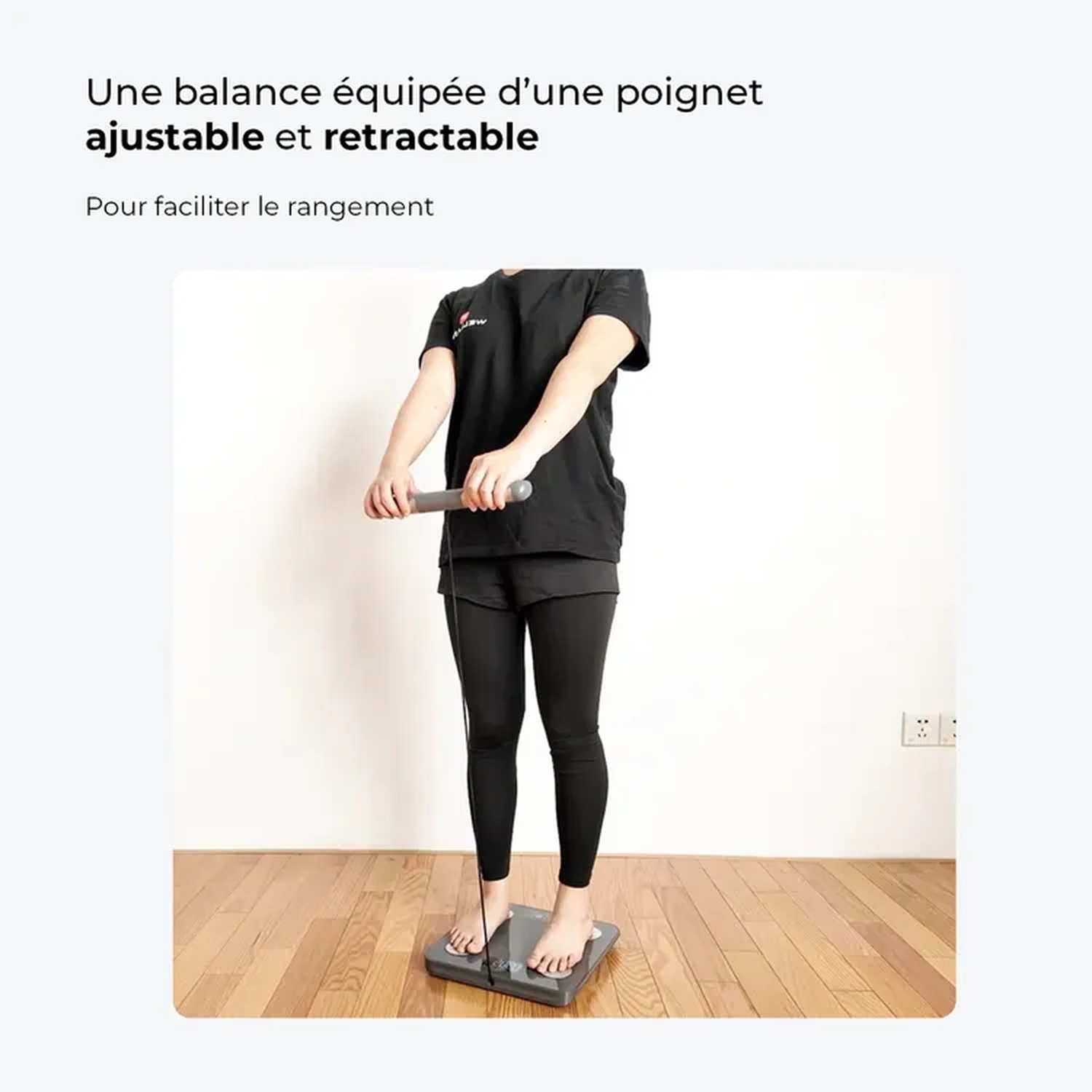 Een persoon staat op een weegschaal en houdt een handvat vast. Ze dragen een zwart shirt, leggings en shorts. De achtergrond is een houten vloer en een witte muur.