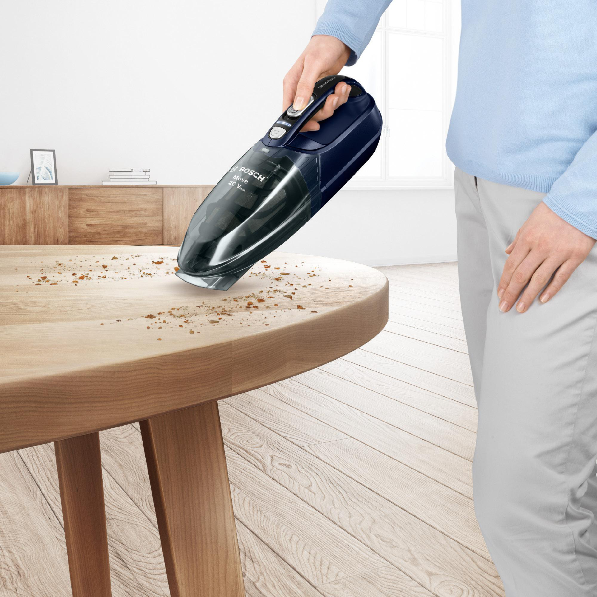 Eine Person verwendet einen kabellosen Staubsauger, um Krümel von einem Holztisch zu entfernen. Der Staubsauger ist schwarz und blau.
