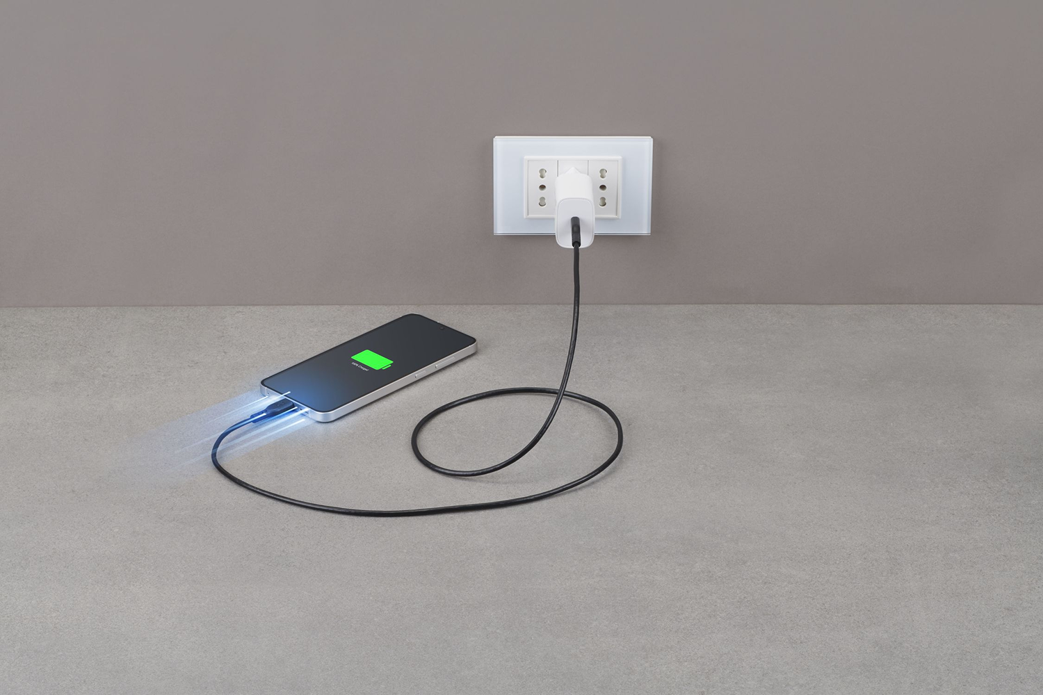 Un teléfono cargándose con un cable conectado. El enchufe está en un enchufe. El teléfono muestra 100% cargado en la pantalla.