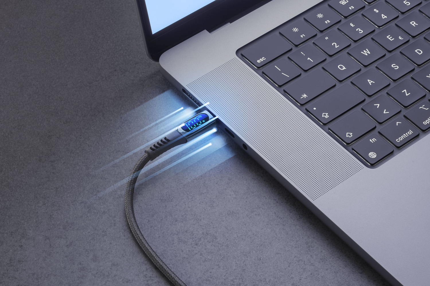 Laptop con cable de carga conectado. El cable muestra una lectura digital. También son visibles el teclado y una superficie gris.