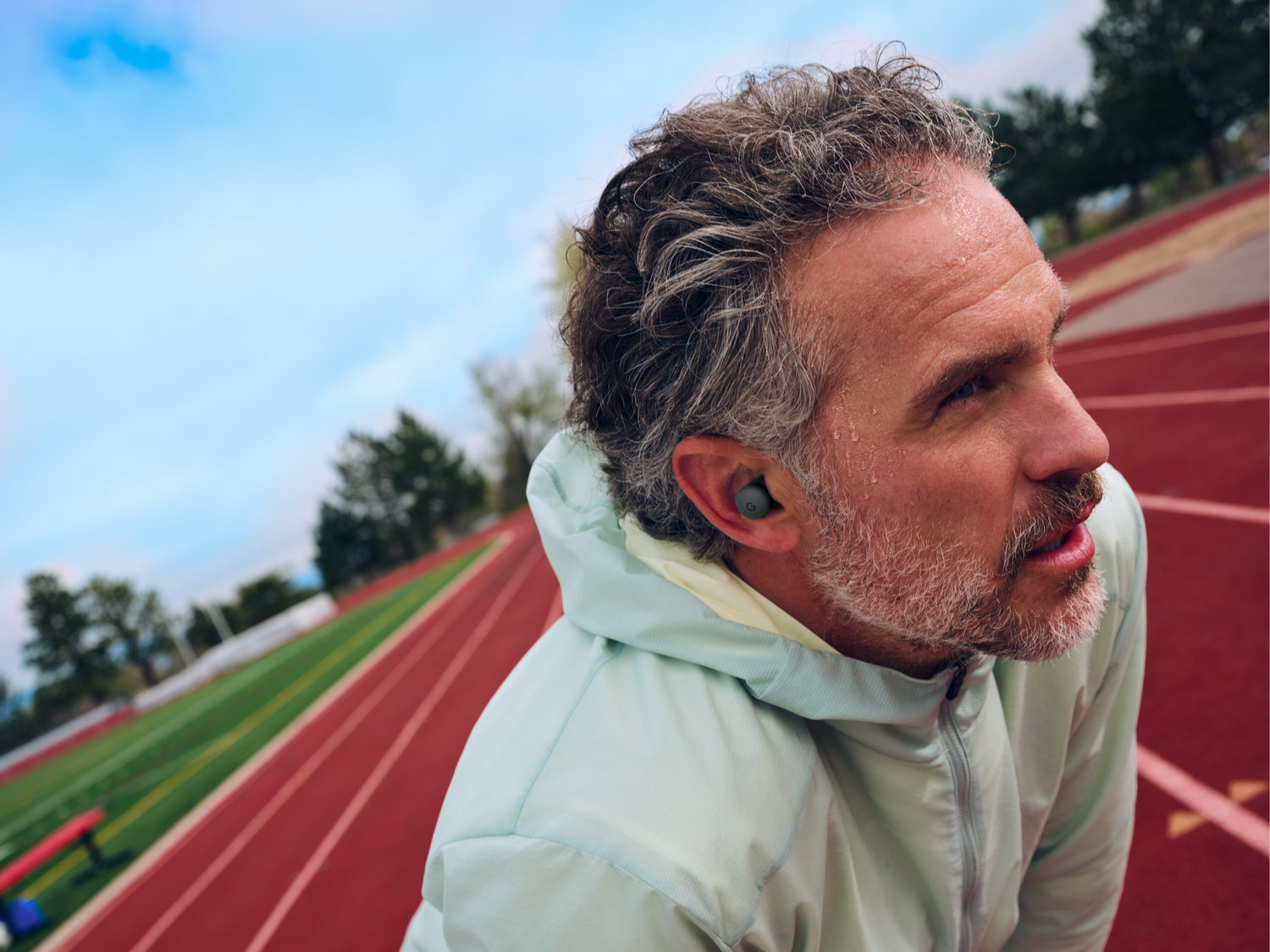 Un hombre con pelo gris y barba, con una chaqueta verde claro, está en una pista de atletismo, mirando hacia un lado. Tiene auriculares.