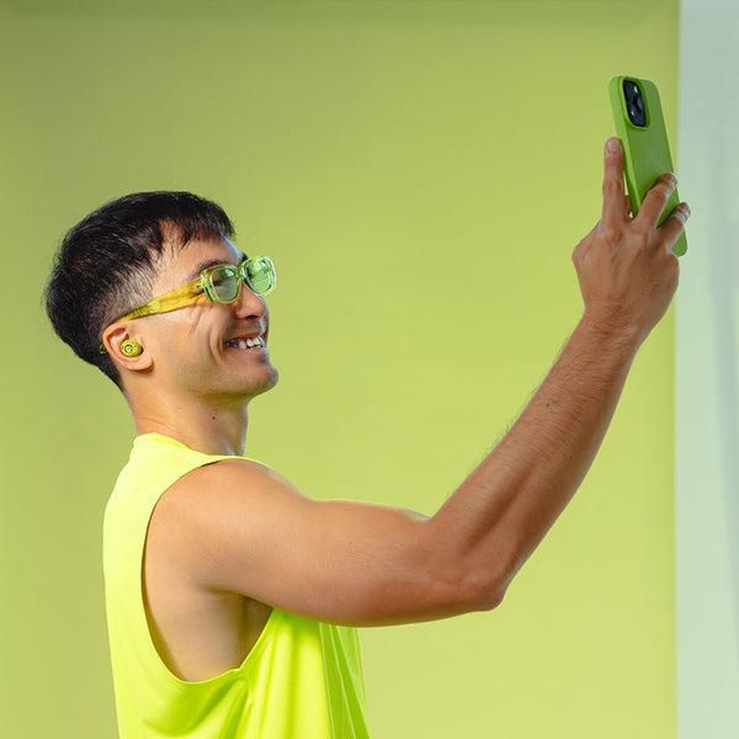 A man in neon green glasses and top takes a selfie with a green phone, smiling. Background is a solid light green.