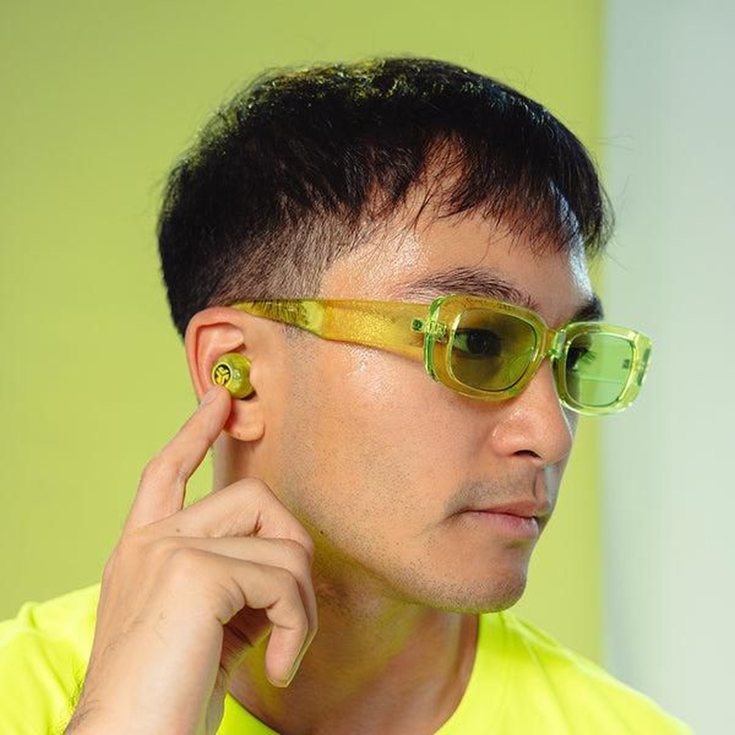 A man with glasses and earbuds. He is touching the earbud. Background is light green.
