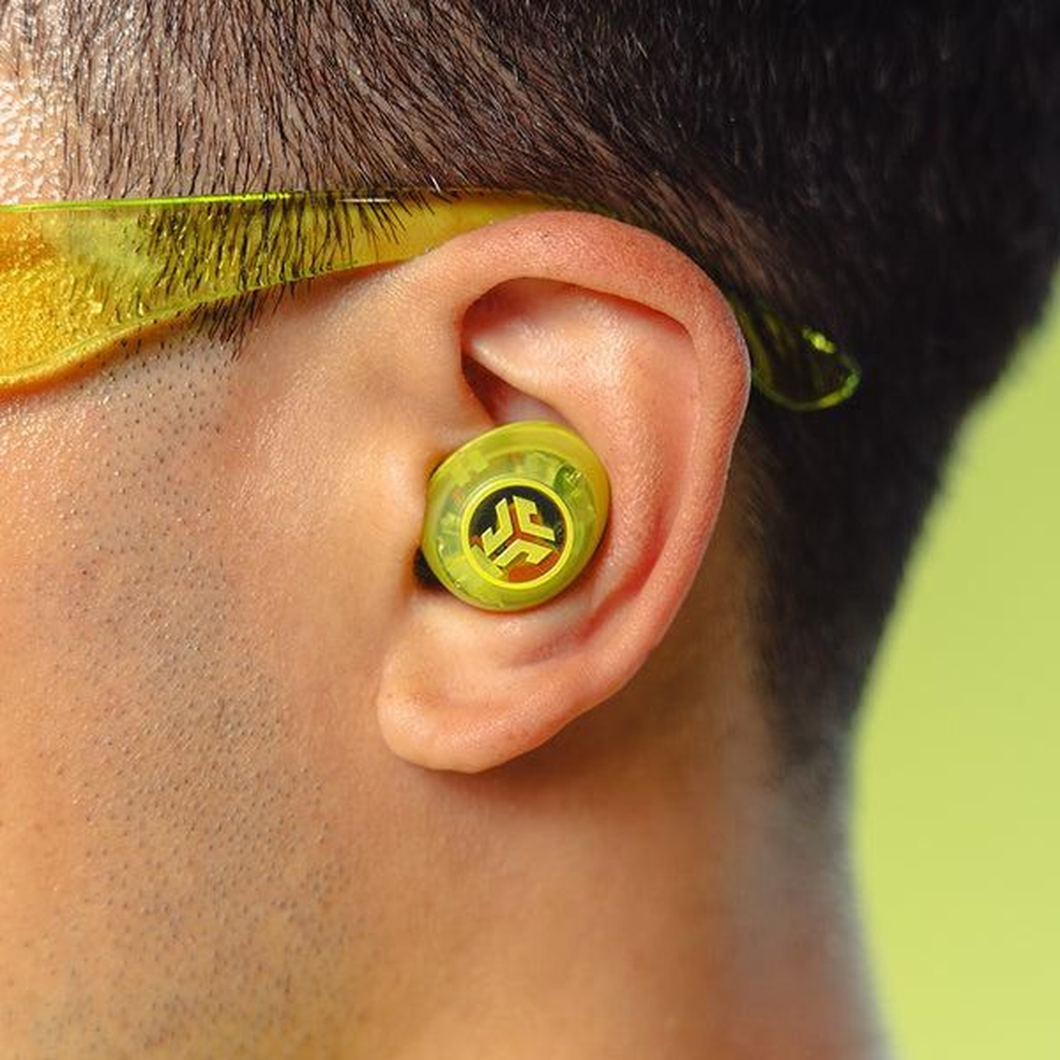 Close-up of a person wearing a yellow earbud. The earbud is inside the ear. Yellow glasses are on the top of the ear.