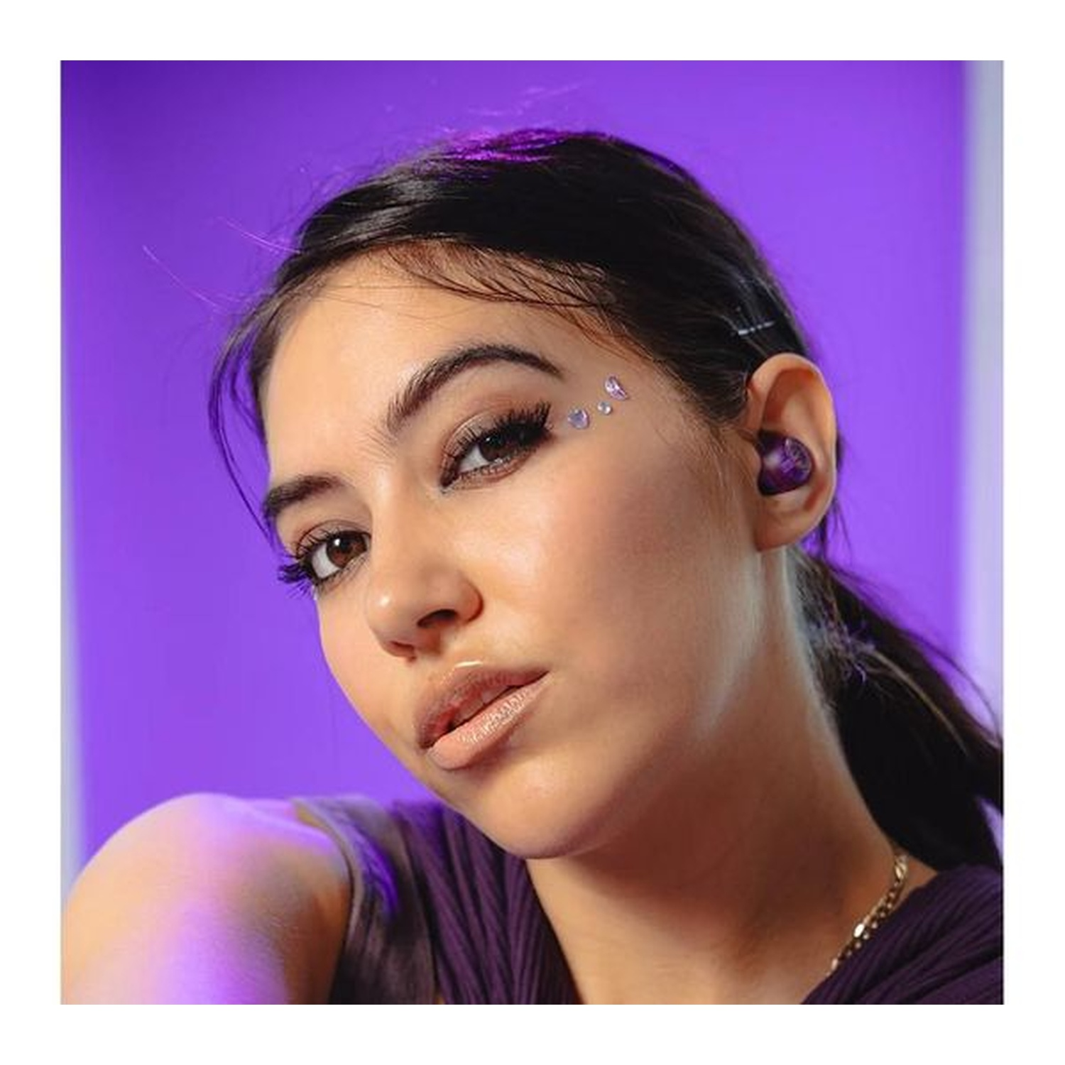 A woman with dark hair and makeup wears a purple top and earbuds. She has gems on her face, a gold necklace, and a purple background.