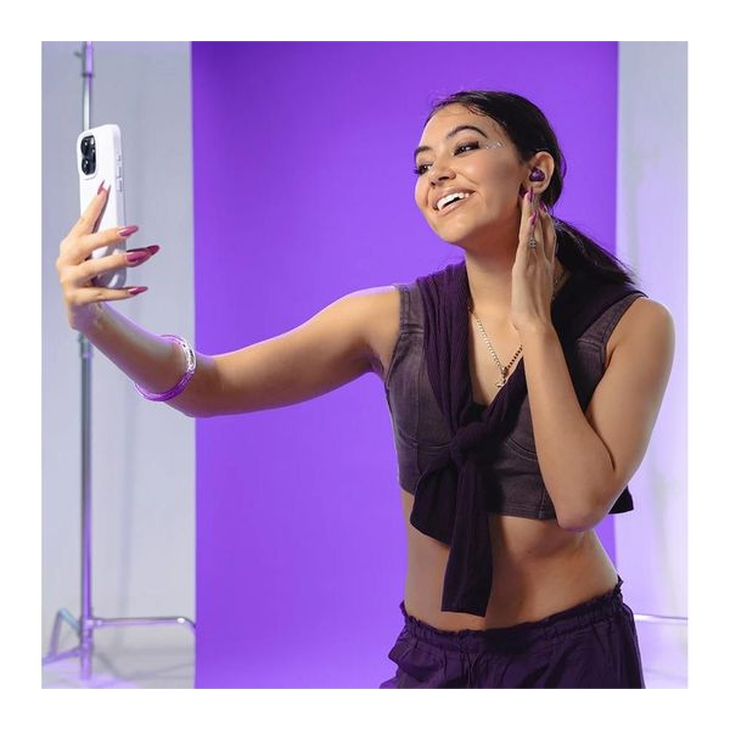A smiling woman takes a selfie with a phone. She wears a purple outfit and has long nails. Purple backdrop in the background.