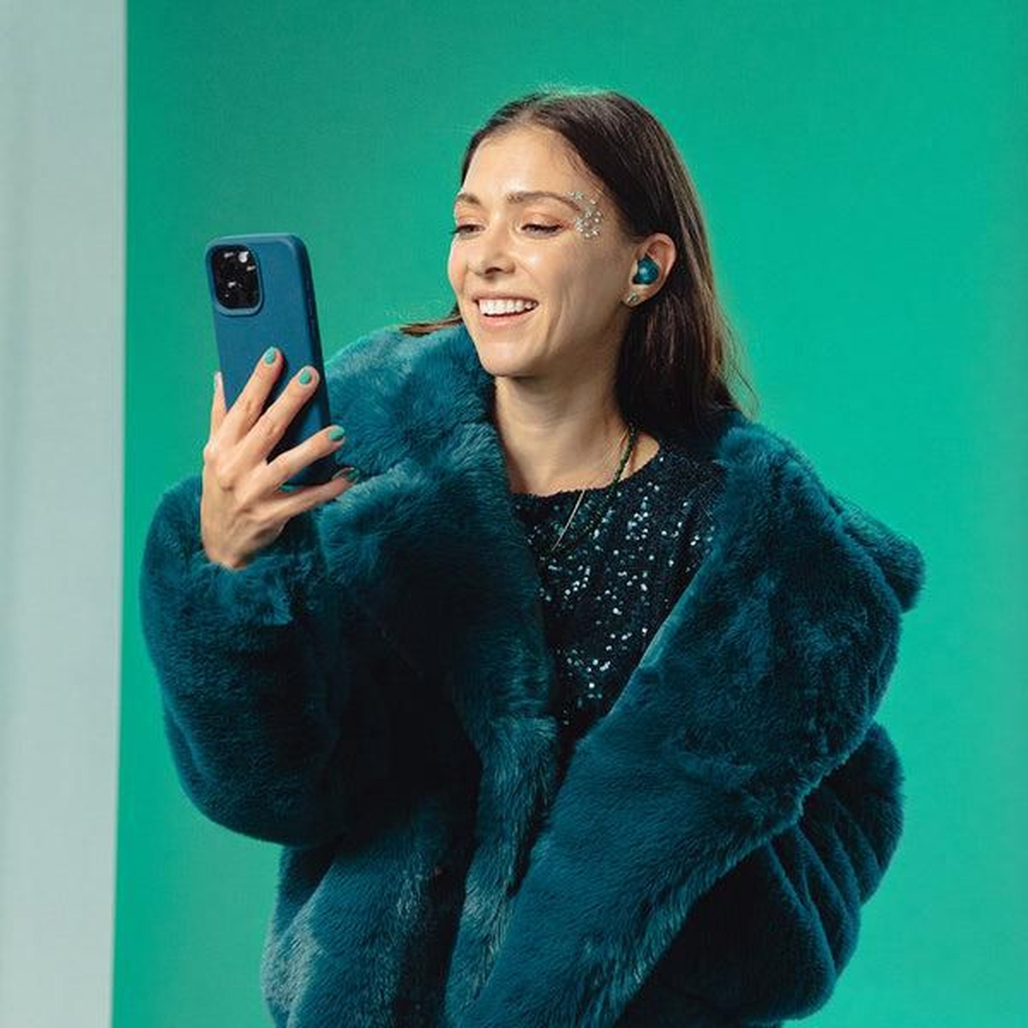 A woman in teal fur coat smiles while holding a phone. She wears earbuds, makeup, and a sequined top against a green backdrop.
