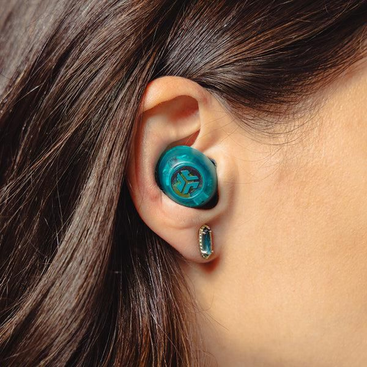 A person's ear with a teal earbud and a matching earring. Brown hair is visible in the background.