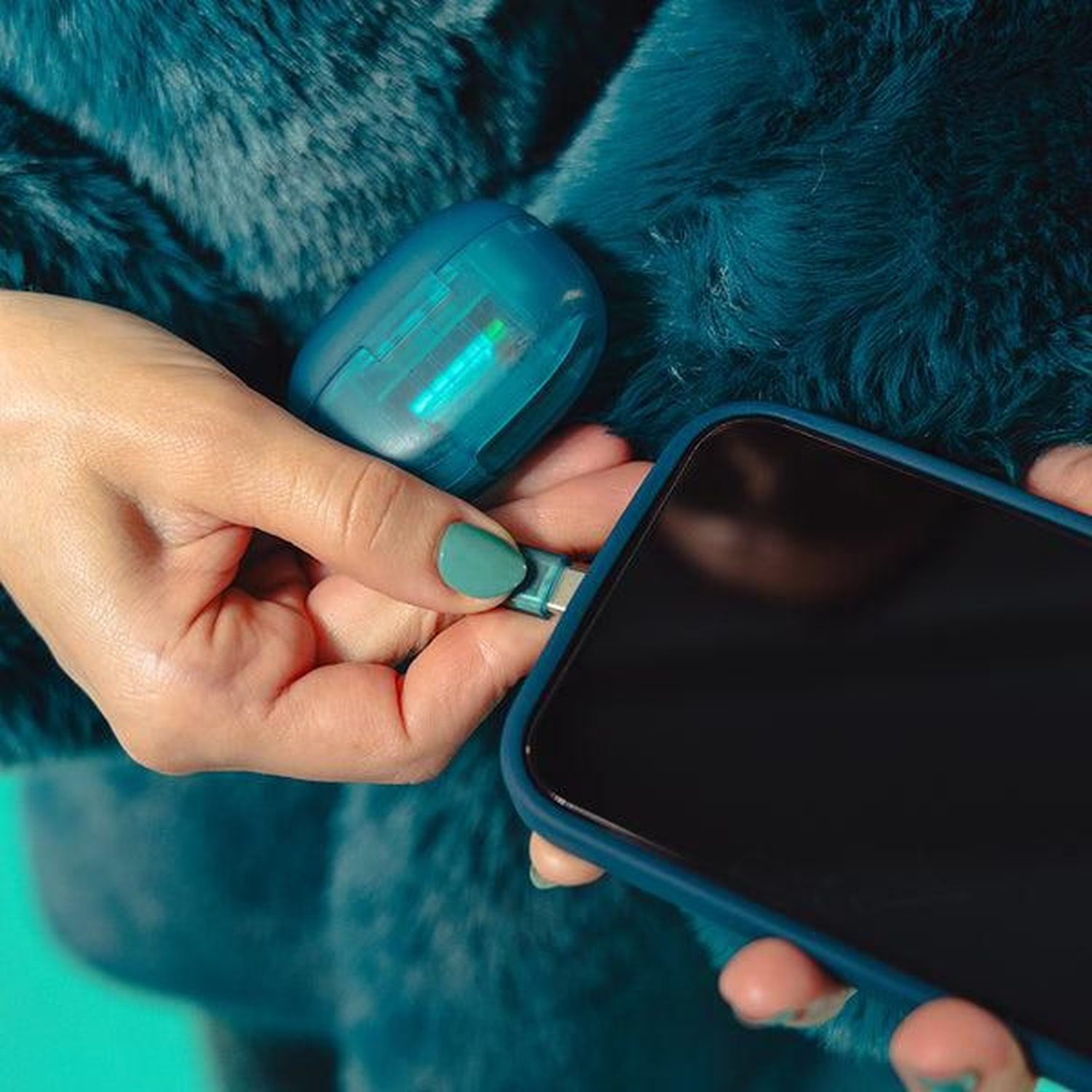 A person holds a blue phone and a blue charging device, connecting them. The background is a teal, fluffy fabric.