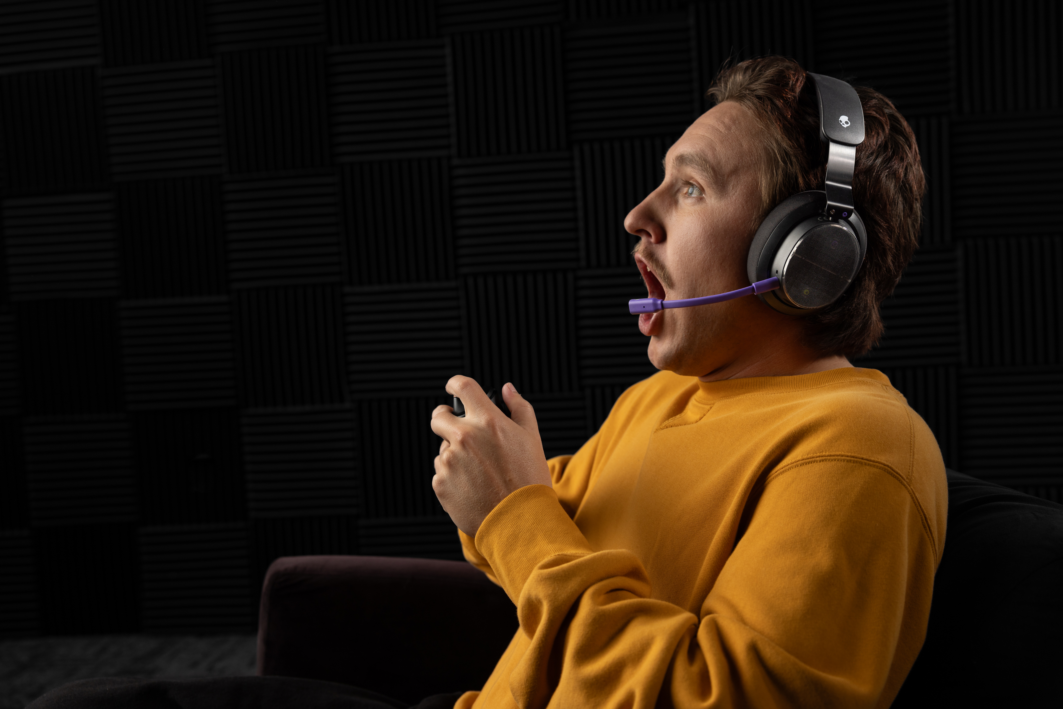 Ein Mann mit Headset und Controller spielt. Er trägt ein gelbes Hemd und hat den Mund offen. Der Hintergrund hat ein schwarzes Muster.