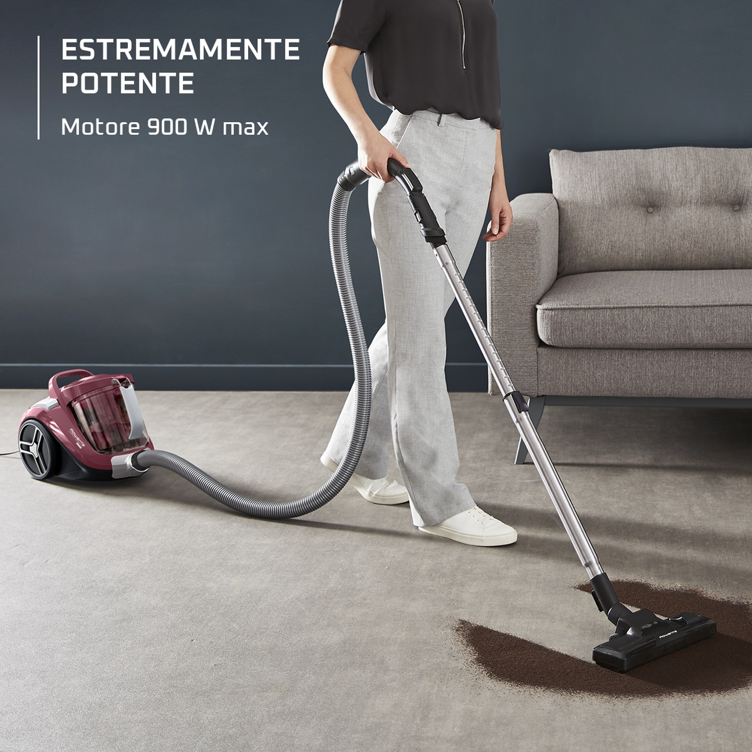 Una persona passa l'aspirapolvere su un tappeto con un aspirapolvere bordeaux. La scena include un divano e il testo 'Estremamente potente, motore 900 W max'.