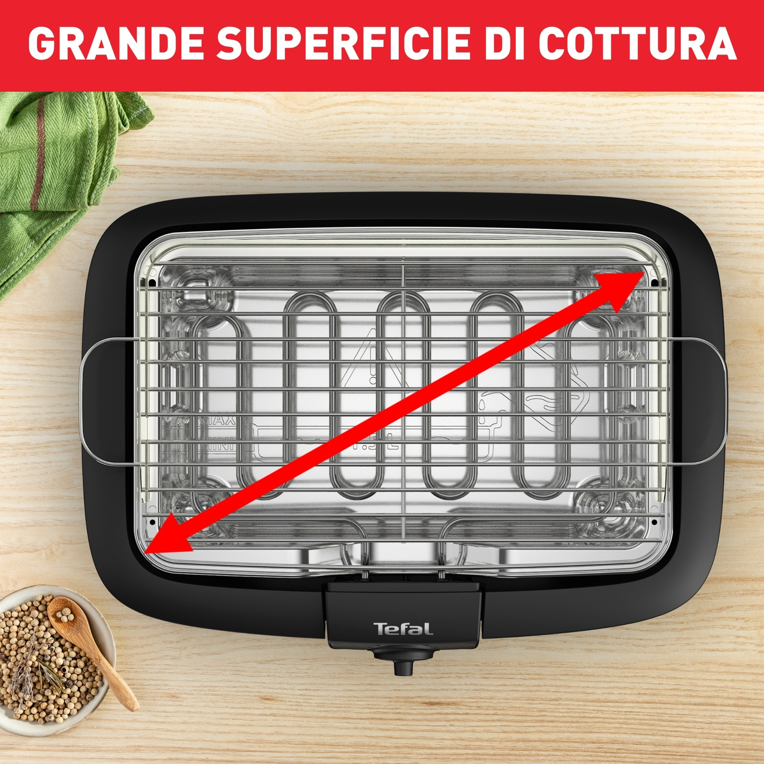 Griglia elettrica con ampia superficie di cottura. Freccia rossa indica l'area di cottura. Spezie in una ciotola e un panno verde di lato.