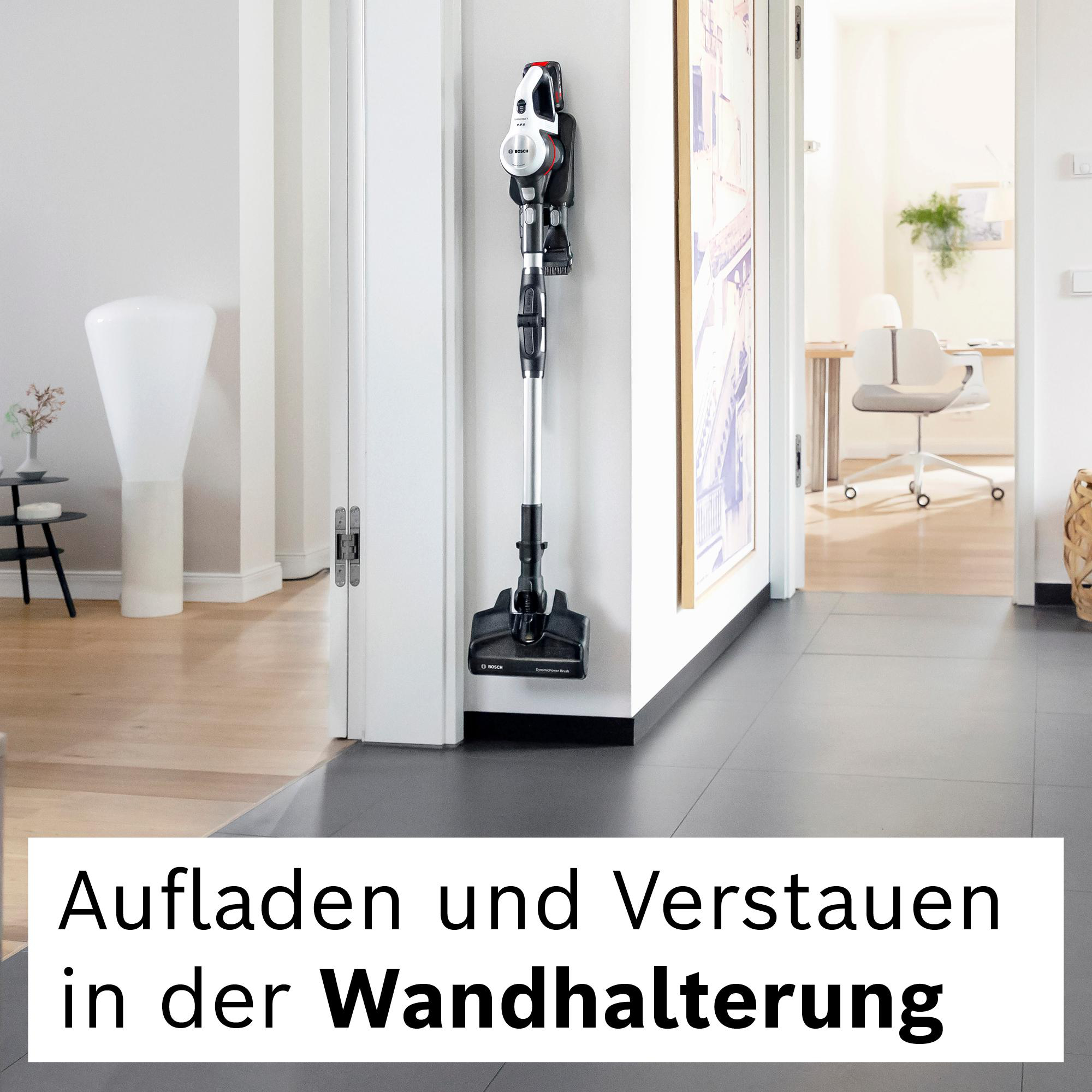 Ein kabelloser Staubsauger ist an einer Wand montiert. Im Hintergrund sind eine Tür und ein Flur mit Schreibtisch und Stuhl zu sehen.