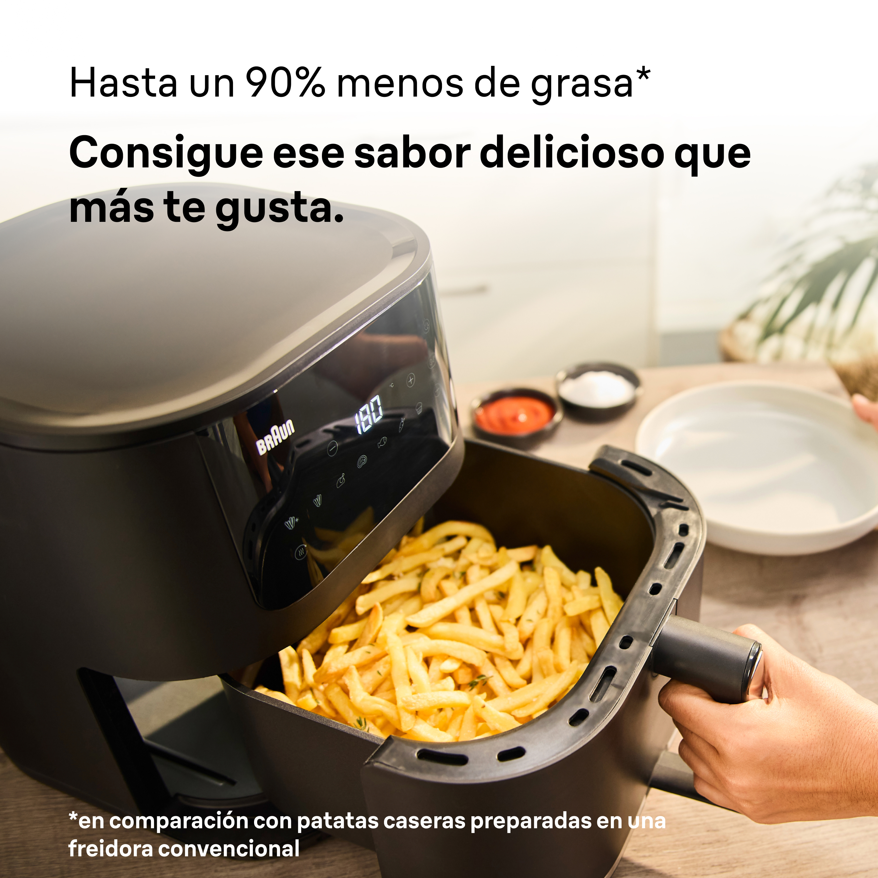 Una freidora con patatas fritas dentro, una mano sostiene la cesta. Un plato y condimentos están sobre la mesa de madera.