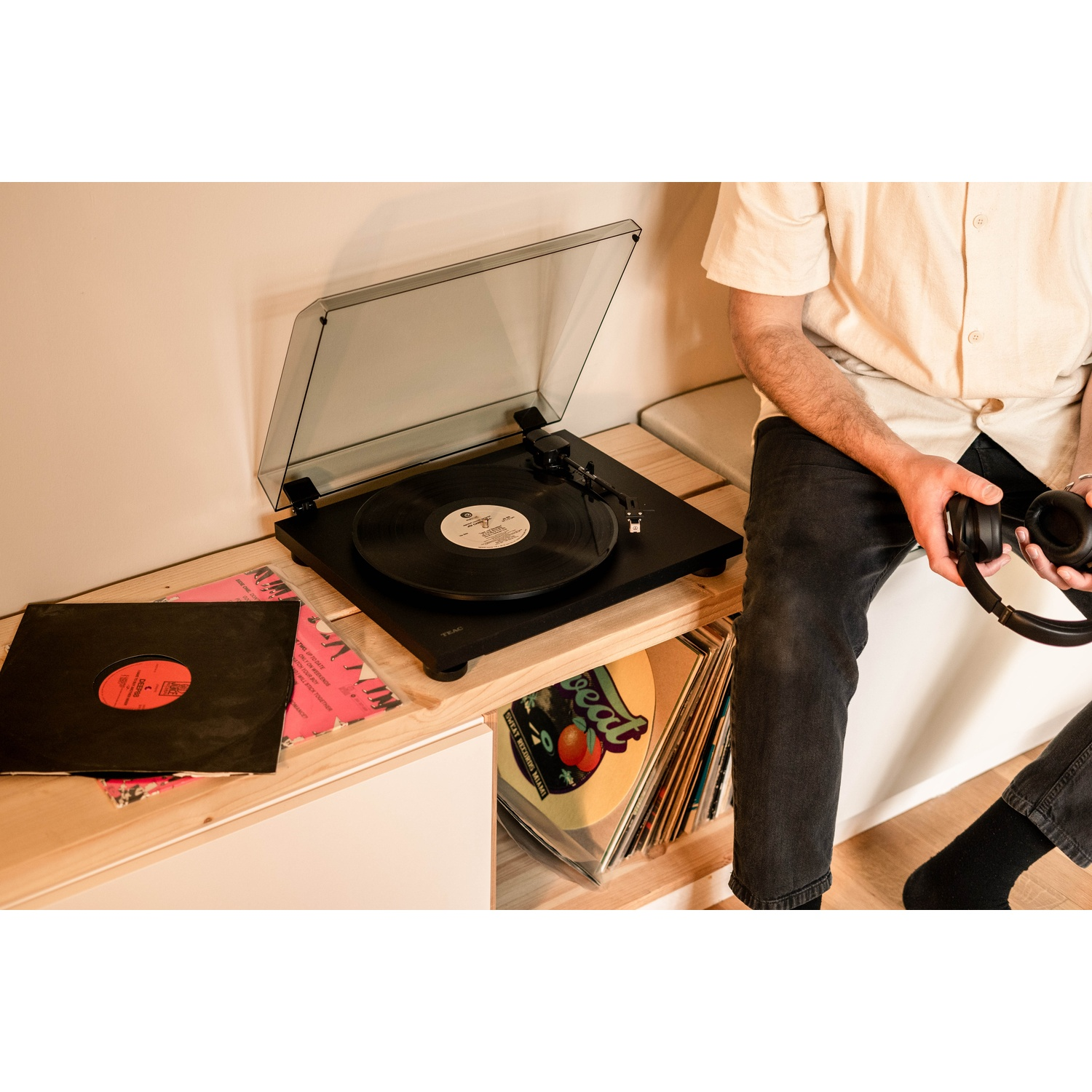 Una persona è seduta vicino a un giradischi. Cuffie in mano. I dischi sono sullo scaffale. Un disco in vinile sta suonando. La persona indossa jeans scuri.