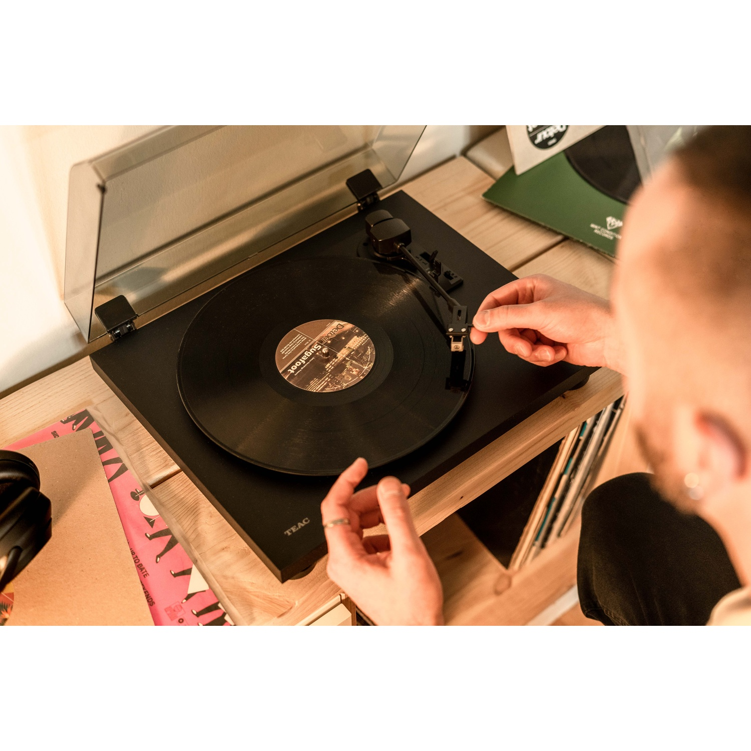 Una persona sta usando un giradischi TEAC nero. Un disco in vinile è sul giradischi. Sono visibili altri dischi e una mensola di legno.