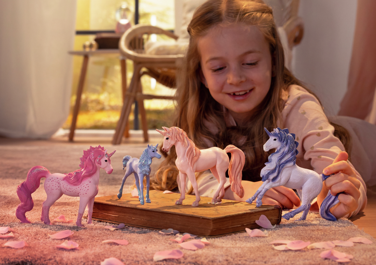 Ein Mädchen lächelt Spielzeug-Einhörner an. Rosa Blütenblätter und ein Teppich im Vordergrund, Tisch und Stuhl im Hintergrund.