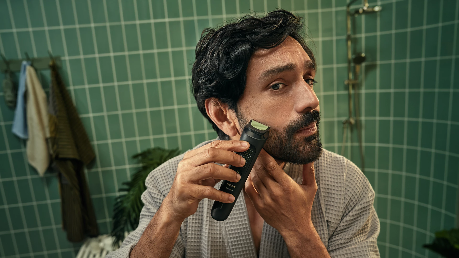 Ein Mann rasiert seinen Bart mit einem schwarzen Philips Trimmer in einem Badezimmer. Der Hintergrund hat grüne Fliesen, einen Duschkopf und Handtücher.