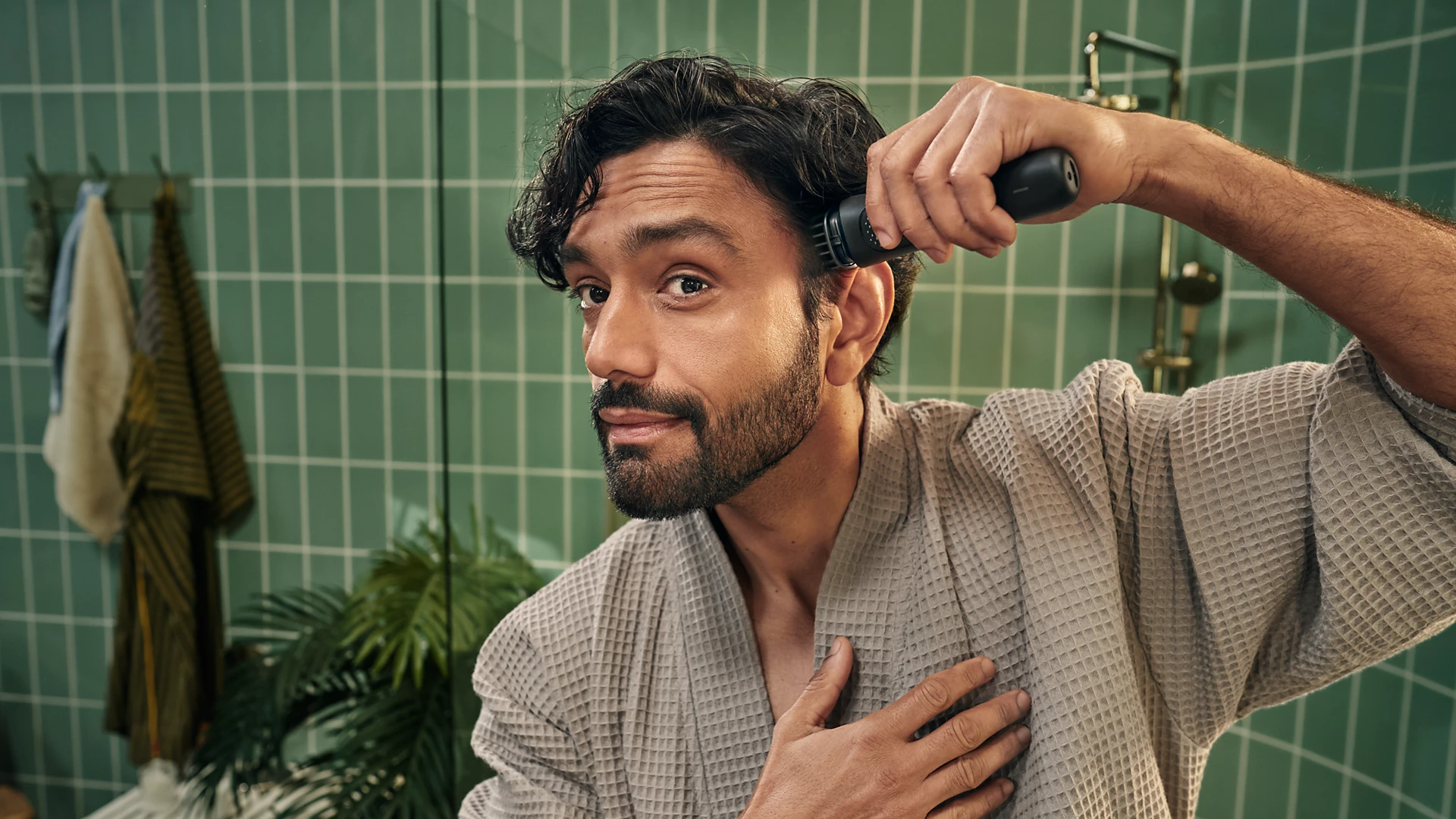Ein Mann rasiert sich im Badezimmer mit einem Trimmer den Kopf. Er trägt einen Bademantel. Grüne Fliesen und Handtücher sind im Hintergrund.
