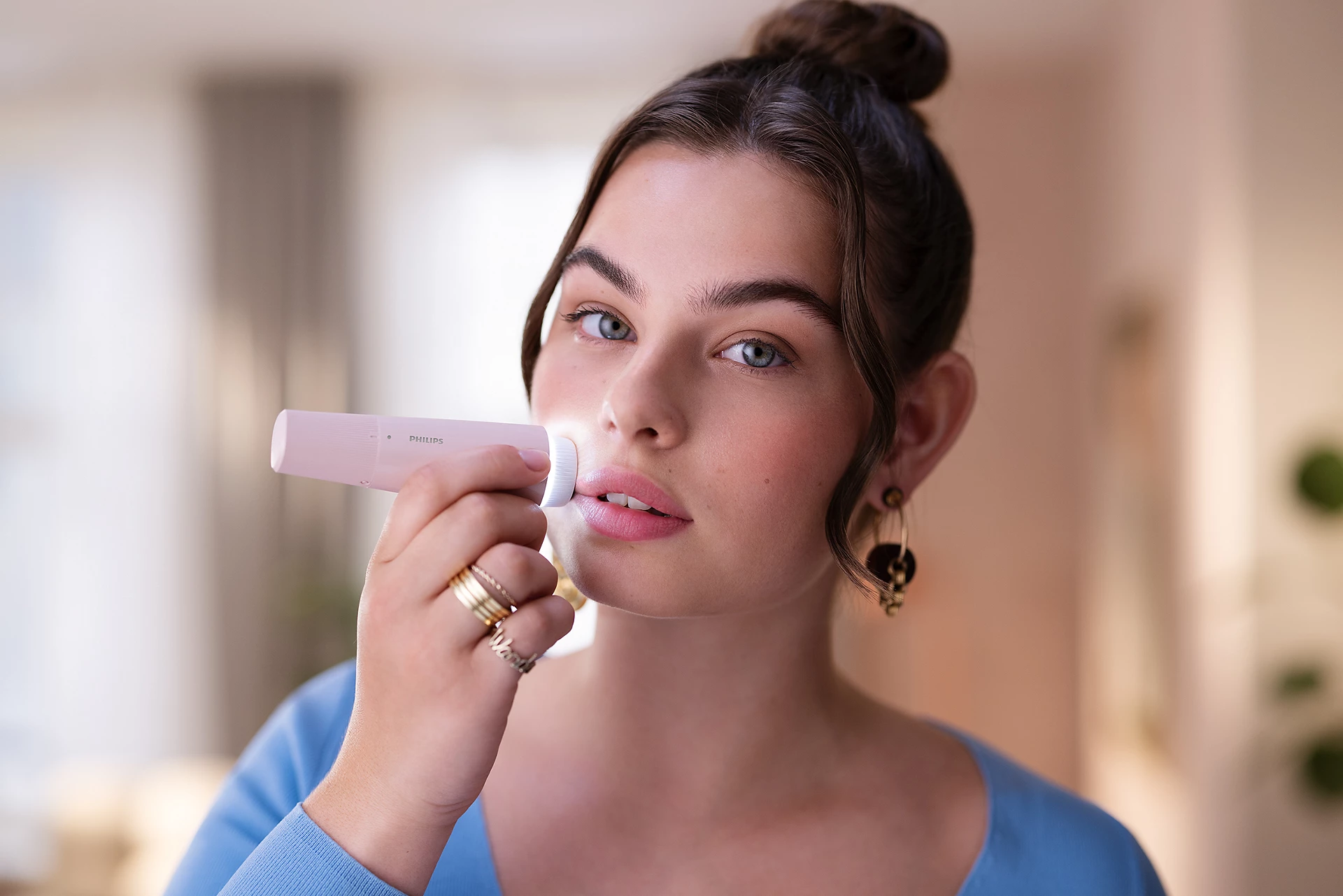 Una donna usa un dispositivo per la pulizia del viso Philips rosa. Ha la pelle chiara, gli occhi azzurri e i capelli raccolti in uno chignon.