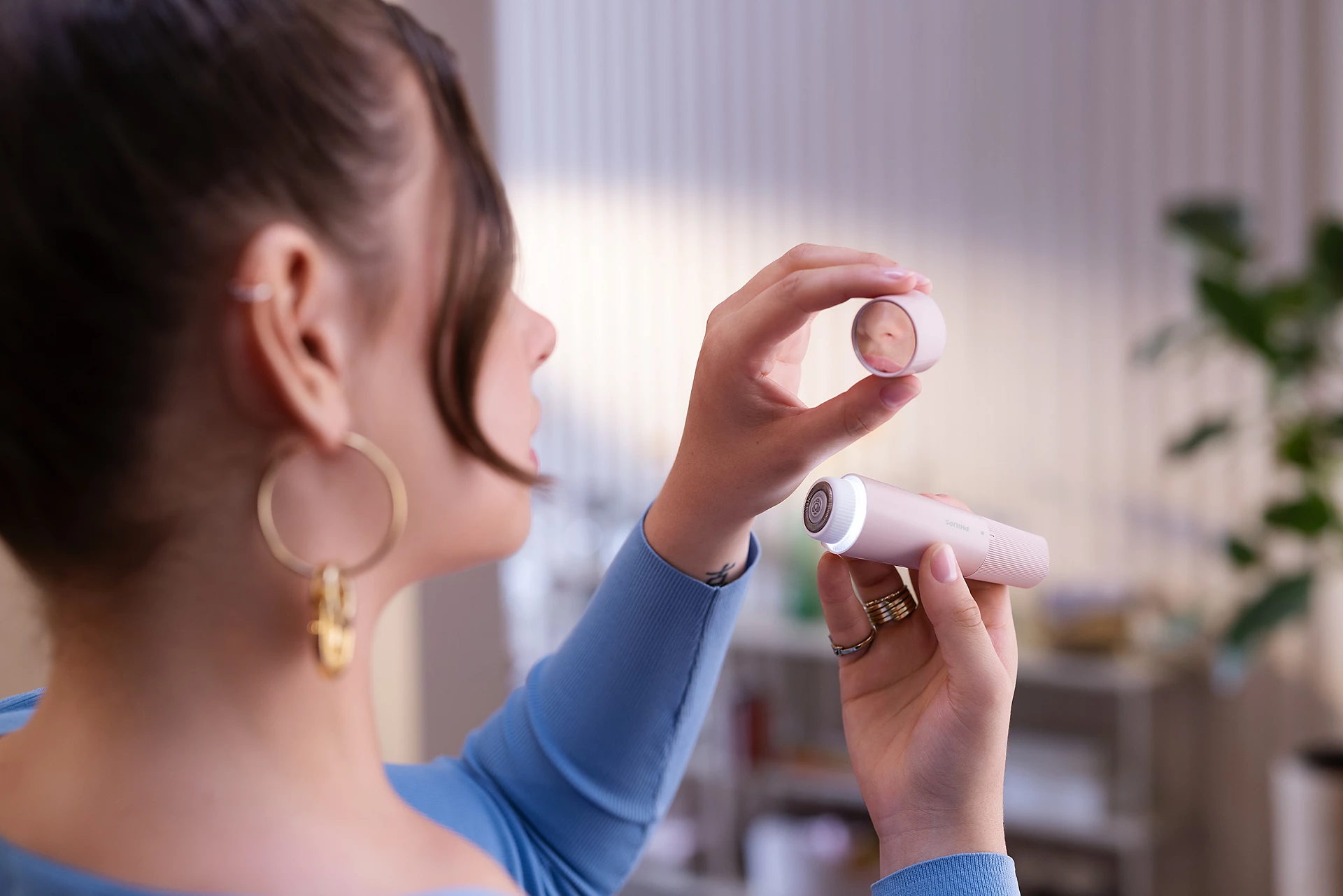 Una donna tiene in mano un dispositivo per la cura del corpo rosa e il suo tappo, guardando il riflesso. Indossa orecchini d'oro e un maglione blu.