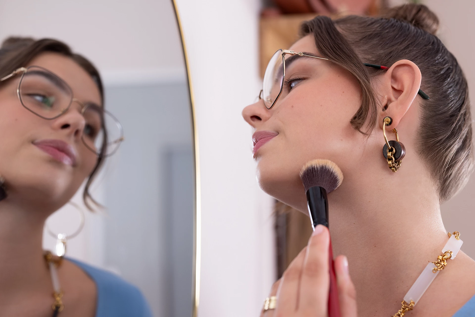 Donna che si trucca davanti a uno specchio. Indossa occhiali, orecchini e una collana. Focus sull'azione.