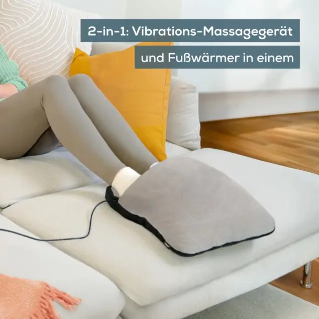 Die Beine einer Person liegen auf einer weißen Couch mit einem Fußmassagegerät. Das Massagegerät ist grau und schwarz. Im Hintergrund ist ein gelbes Kissen.