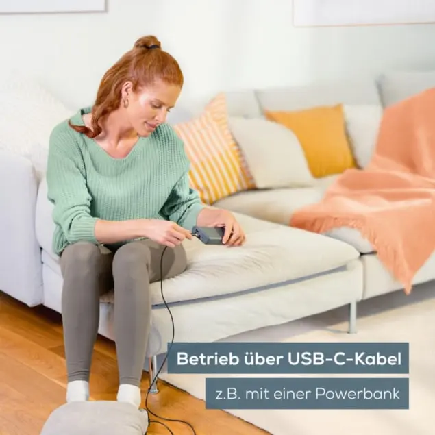 Eine Frau sitzt und verbindet ein Kabel mit einem Gerät auf einem Sofa. Eine Fußwärmer liegt am Boden. Text unten sagt 'USB-C-Kabel'.