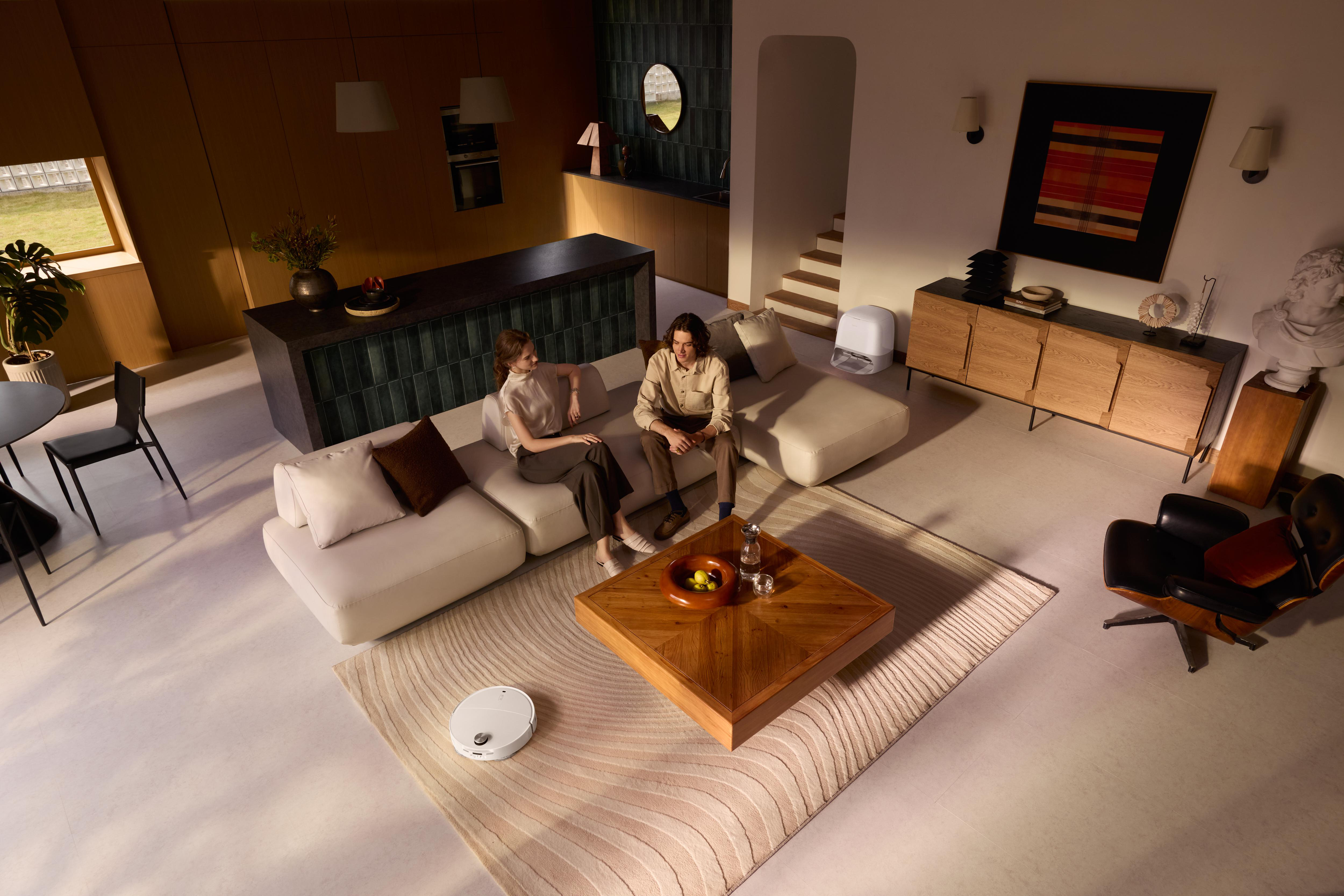 Zwei Personen sitzen auf einem weißen Sofa. Ein Roboter-Staubsauger steht auf dem Boden. Ein Holztisch steht vor ihnen.