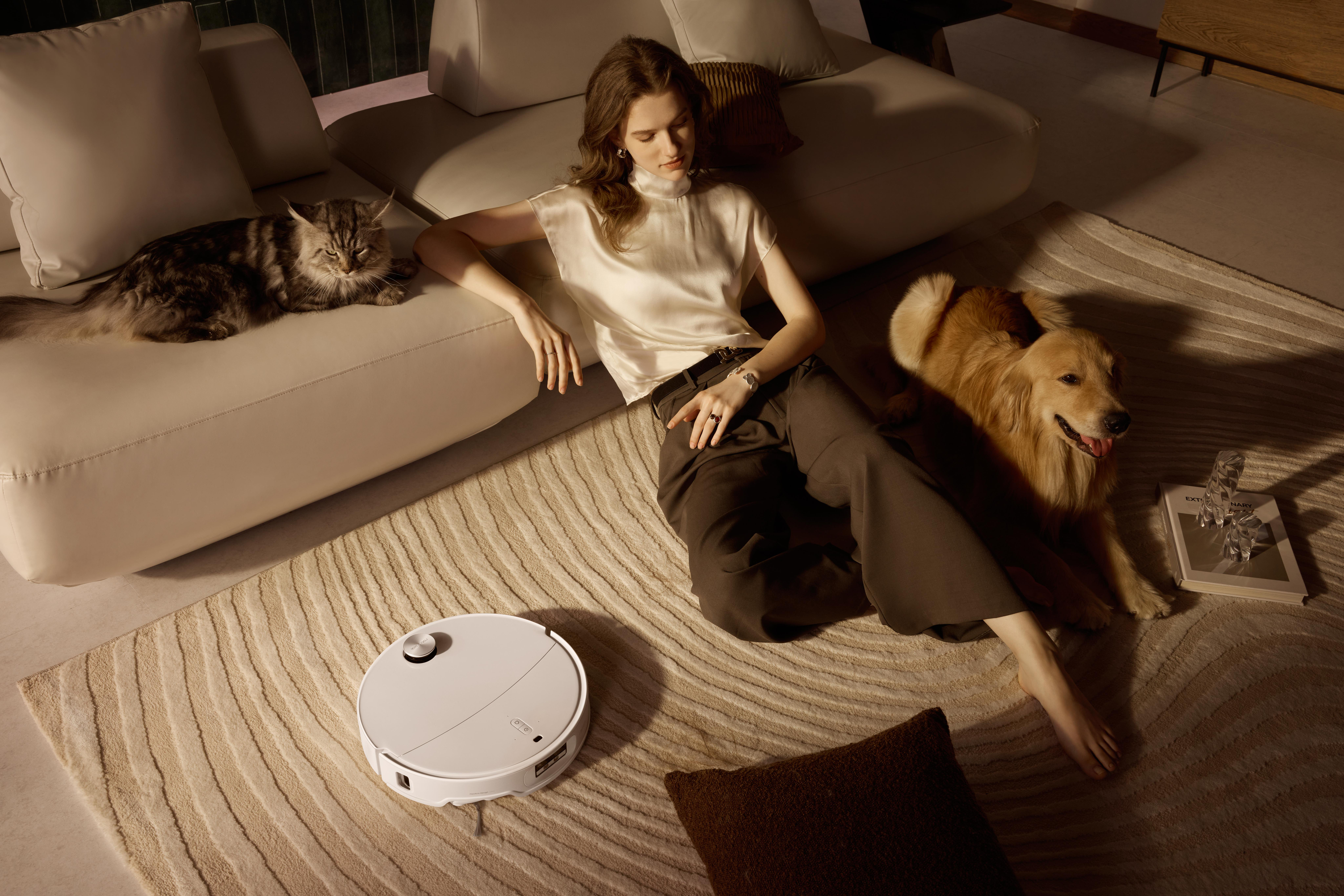 Eine Frau entspannt sich mit Katze und Hund auf einem Teppich. Ein Roboterstaubsauger steht daneben. Die Szene ist ein Wohnzimmer.