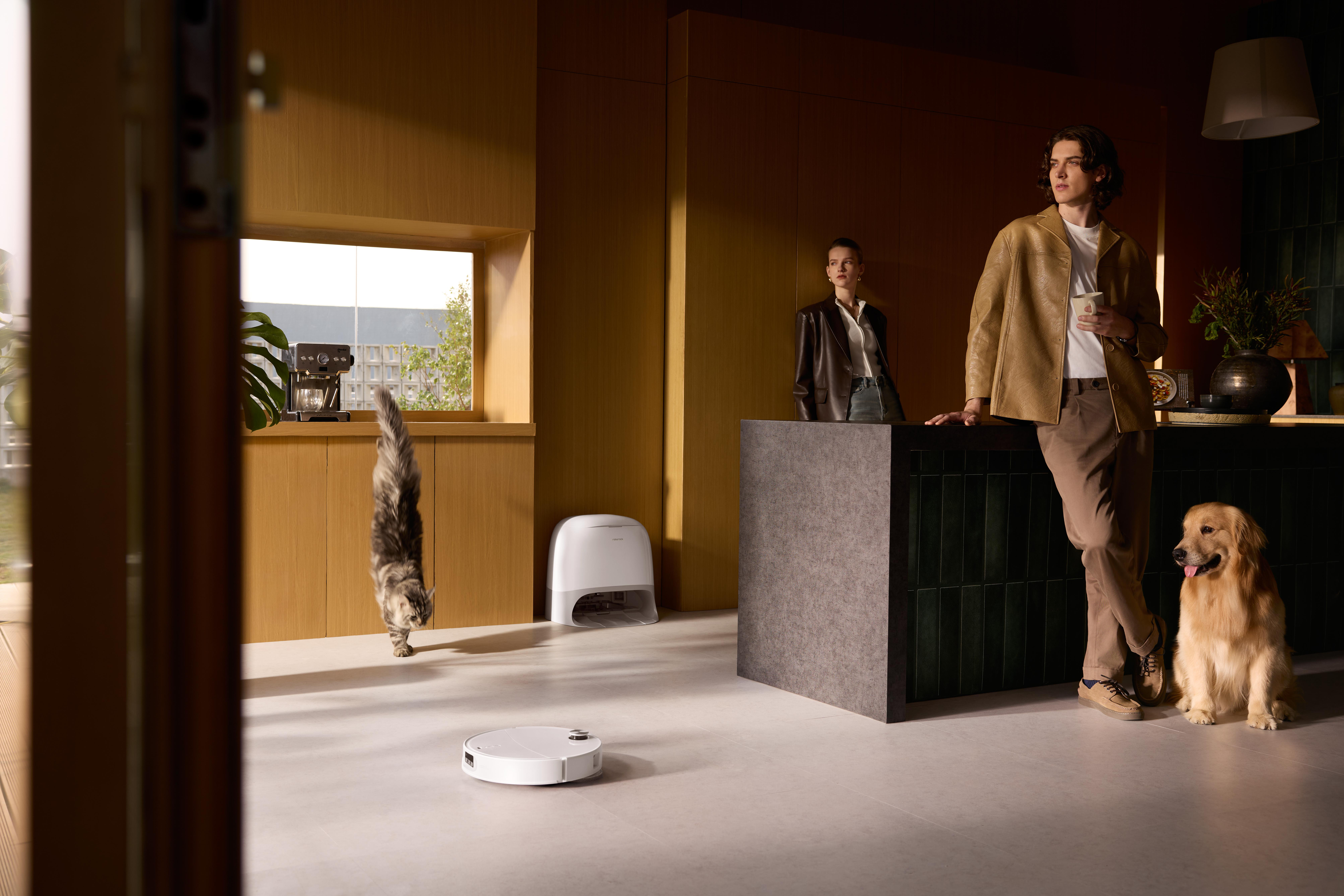 Eine moderne Küchenszene mit Person, Katze, Hund und Roboterstaubsauger. Mit Fenster, Arbeitsplatte und Holzschränken.