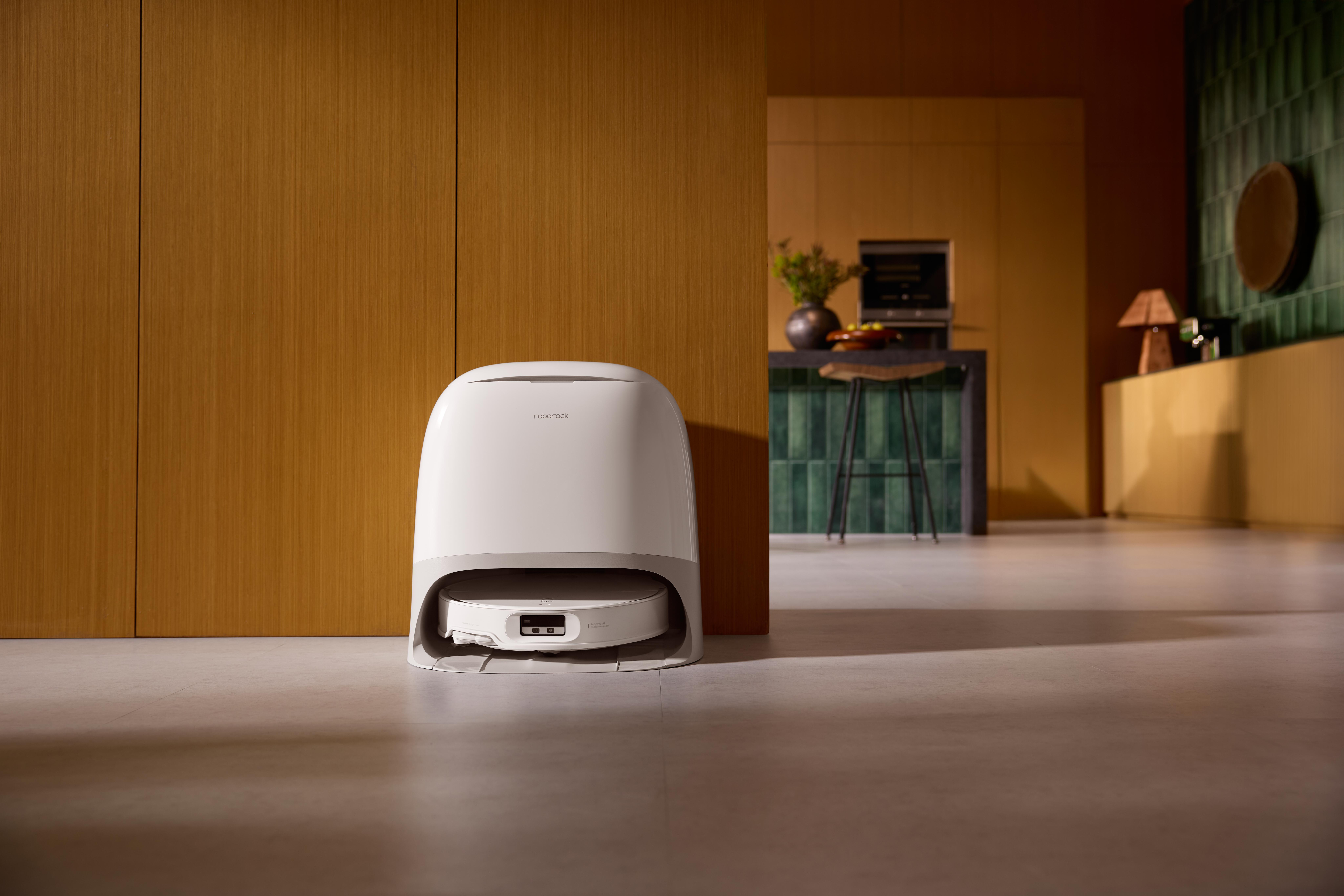 Ein weiß-grauer Roboter-Staubsauger steht in einem modernen Raum. Holzwand und Küche im Hintergrund.