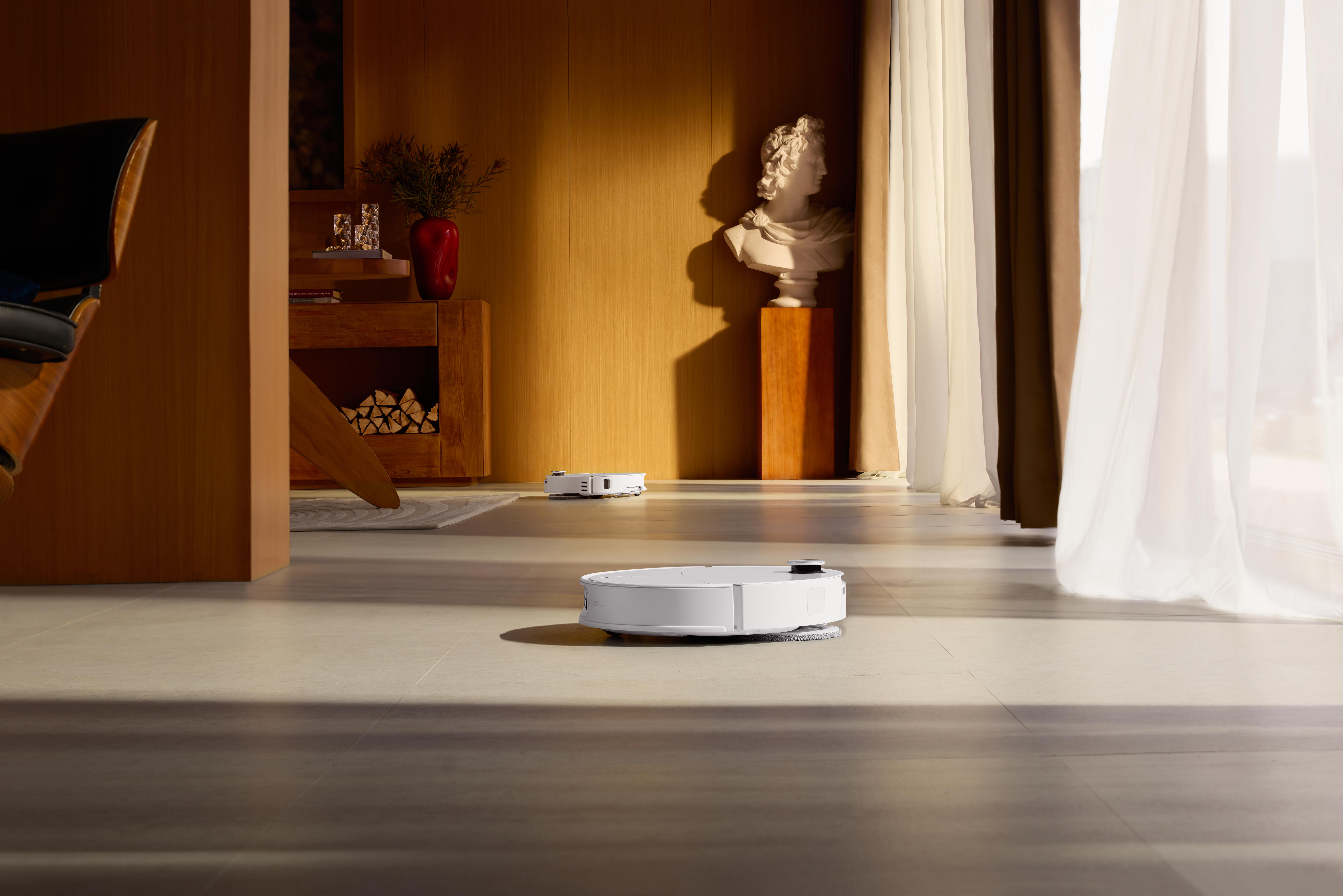 Ein modernes Wohnzimmer mit zwei Roboterstaubsaugern, die den Boden reinigen. Eine Büste, Holzmöbel und transparente Vorhänge sind sichtbar.