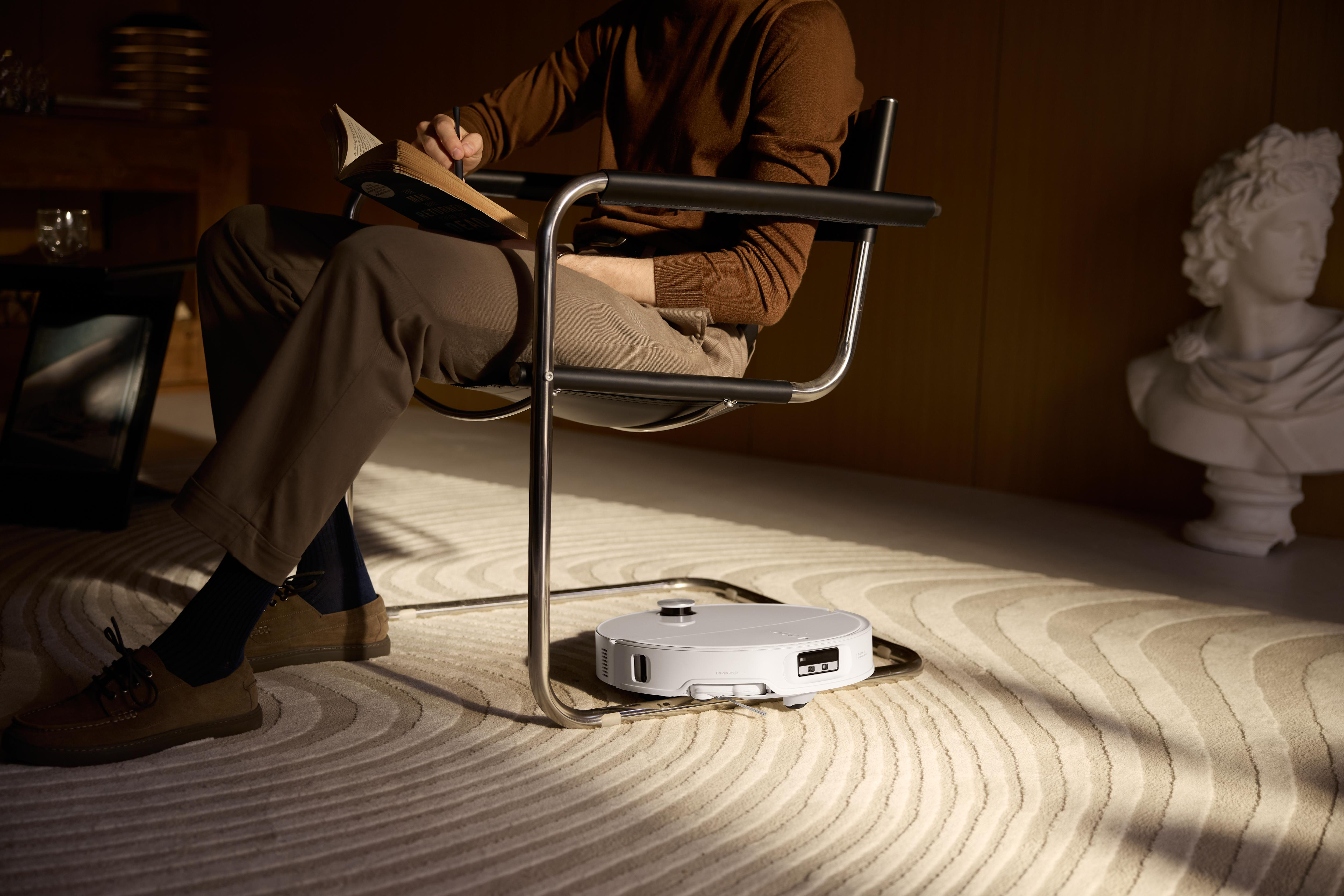 Ein Mann liest auf einem Stuhl, während ein Roboter-Staubsauger auf einem Teppich arbeitet. Eine Büste und ein Bilderrahmen sind im Hintergrund.