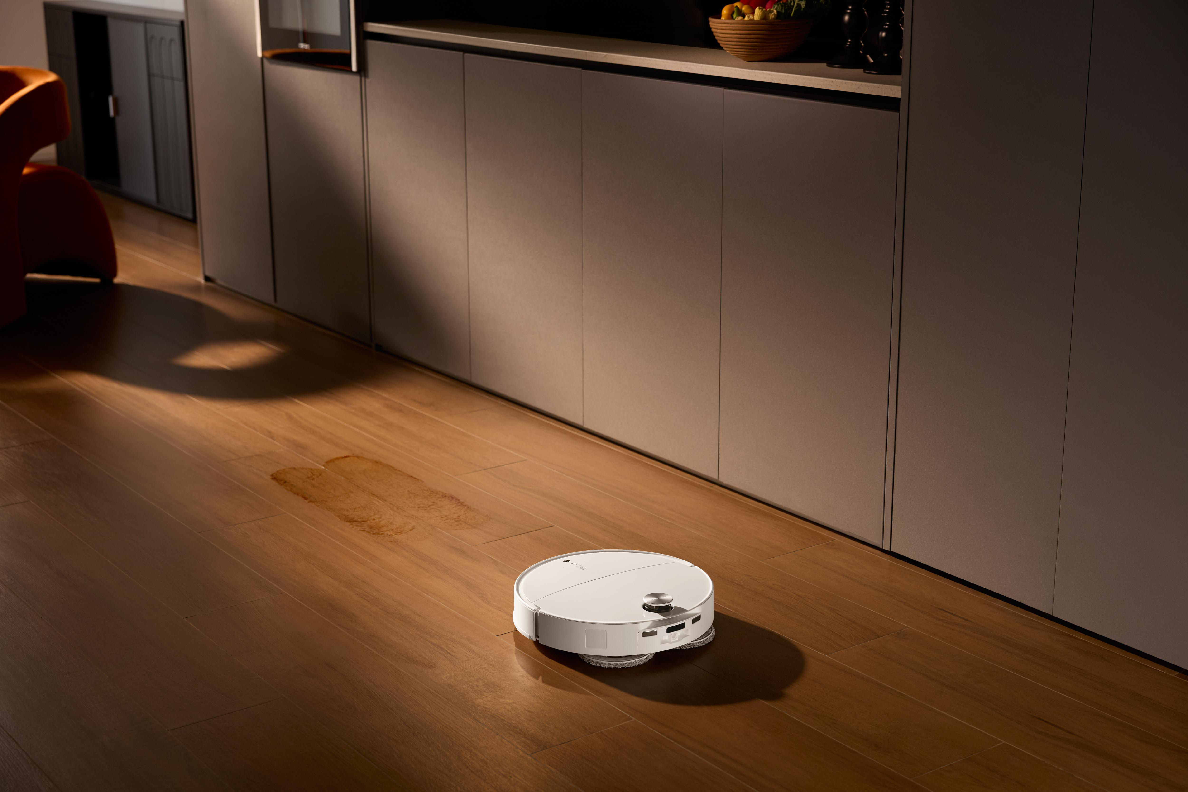 Ein weißer Roboter-Staubsauger auf einem Holzboden. Ein nasser Fleck ist auf dem Boden. Schränke sind im Hintergrund.